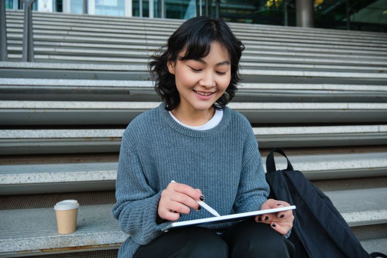 Smiling girl, graphic designer, using digital tablet and pen tool to draw, does home assignment for university, sits on stairs